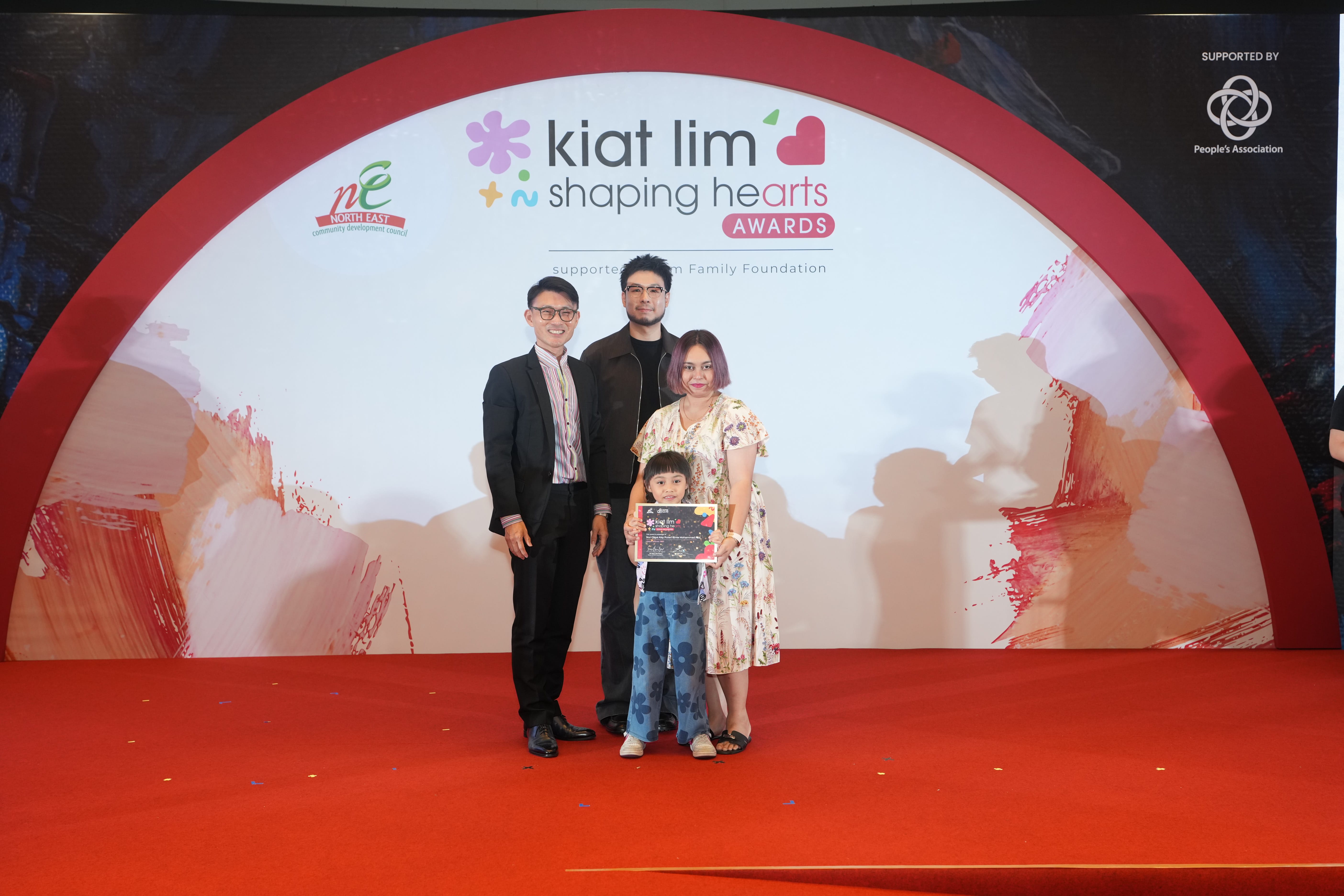 Award recipient standing on stage with the presenter, holding their certificate and posing for a group photo during the award ceremony, with a backdrop reading ‘Kiat Lim-Shaping Hearts Awards'.